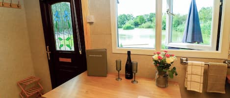 A room with a wooden table, a clock, a bottle of champagne, and a window with a view of greenery.