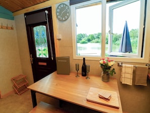 A room with a wooden table, a clock, a bottle of champagne, and a window with a view of greenery. - Moorhen Lodge (Market Rasen)