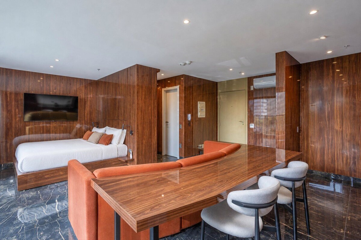 A modern hotel room with a large bed, a wooden dining table, and a TV mounted on the wall.