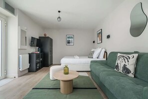Ein modernes Wohnzimmer mit einem grünen Sofa, einem runden Couchtisch, einem weißen Bett und einer schwarzen Kommode.