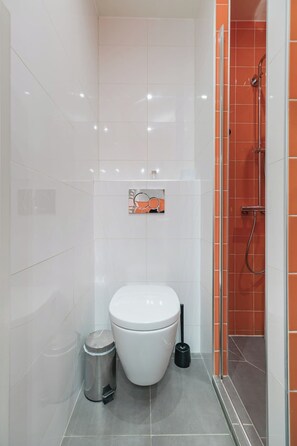Ein modernes Badezimmer mit einer weißen Toilette, einem Mülleimer und einer Duschkabine mit orangefarbenen Fliesen.