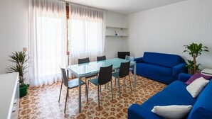 A living room with a glass-top dining table, blue sofa, and a potted plant. - Bright Family Retreat with Terrace, Garden & Beach Nearby (Lido)