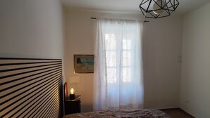 A bedroom with a bed, a nightstand with a lamp, a painting on the wall, and a window with sheer curtains.