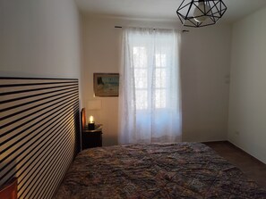 A bedroom with a bed, a nightstand with a lamp, a painting on the wall, and a window with sheer curtains. - Yos 1 Apartment (Vallerano)