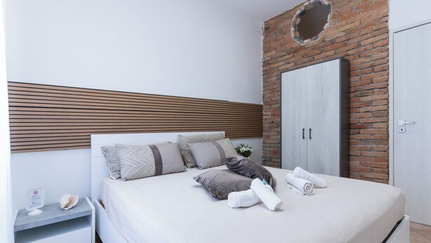 A modern bedroom with a brick wall, a wooden slat headboard, a bed with grey and white bedding, a nightstand, and a wardrobe.