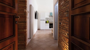 A hallway with a brick wall and wooden doors leading to a kitchen area with white cabinets and a tiled floor.
