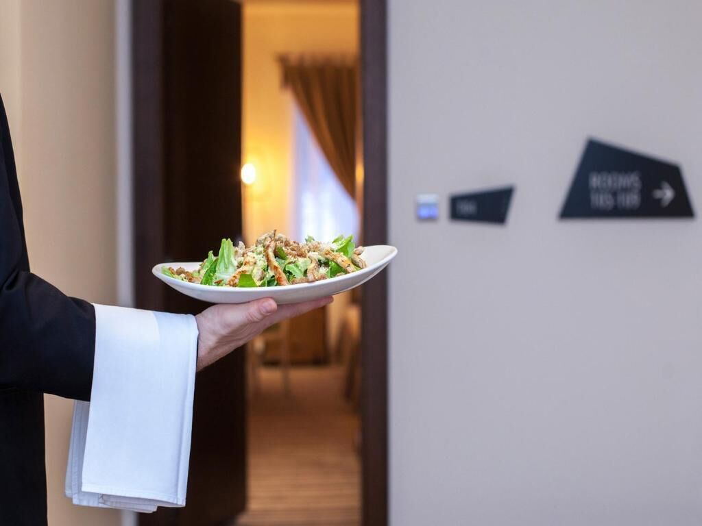 Una persona sosteniendo un plato de comida en un pasillo de hotel con indicaciones.