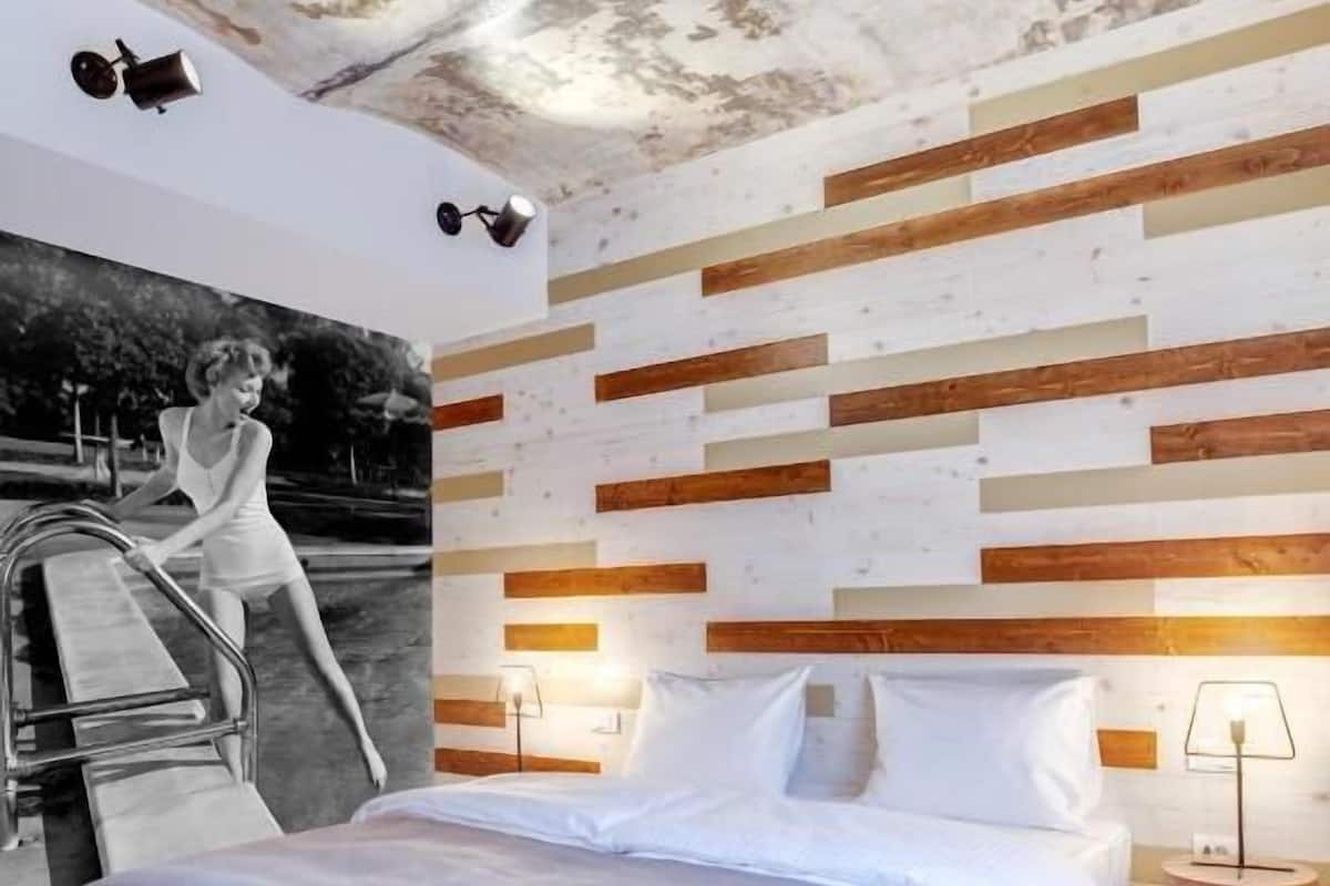 Una habitación de hotel con cama, lámpara de pared, una mesita y una silla.