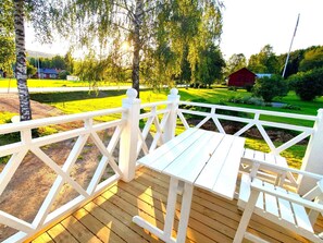 Una terraza de madera con barandas blancas, una mesa y sillas, con vistas a un paisaje verde con árboles y un edificio rojo a lo lejos.
