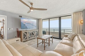 Ein Wohnzimmer mit einer Couch, einem Holztisch, einem Fernseher und einem Balkon mit Meerblick.