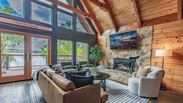 A cozy living room with a stone fireplace, comfortable seating, and large windows offering a view of the outdoors.