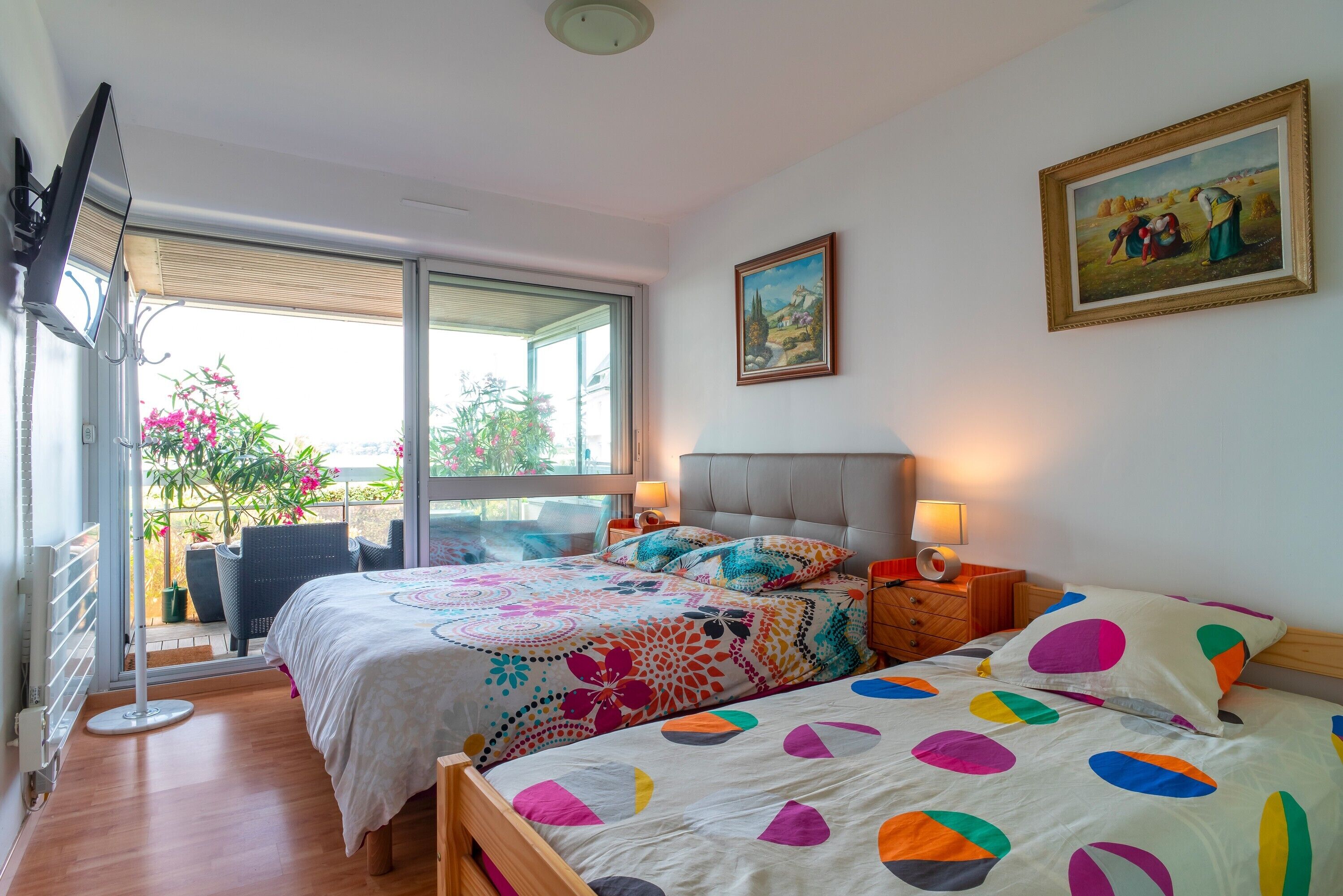 A bedroom with a large bed, a bedside table with a lamp, a painting on the wall, and a view through a sliding door.