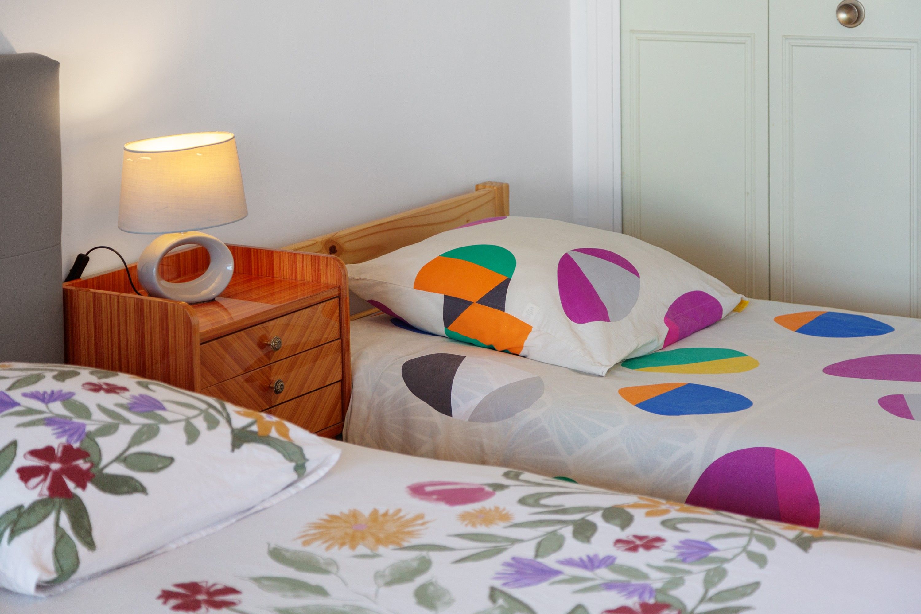 A bedroom with a wooden nightstand, a lamp, a bed with colorful geometric-patterned bedding, and a floral-patterned pillow.
