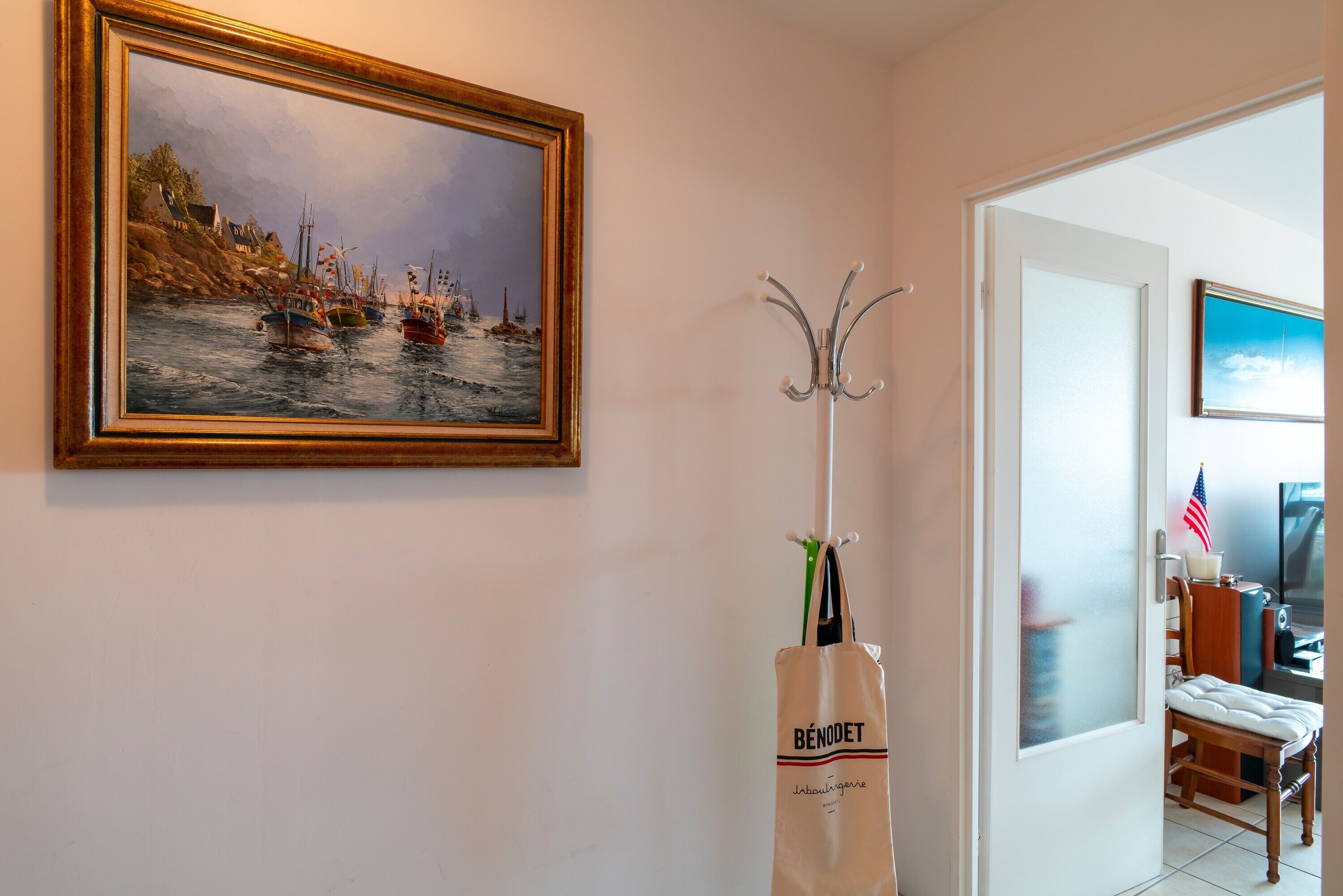 Un couloir avec un tableau encadré représentant des bateaux, un porte-manteau, une porte blanche et une chaise.