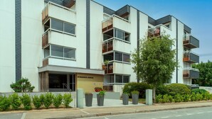 Un moderno edificio residencial de varios pisos con balcones, una entrada visible y plantas en macetas.