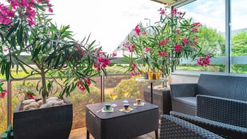 Un espace extérieur aménagé, abrité et doté de mobilier en osier, d’une table avec des boissons et de plantes en fleurs.