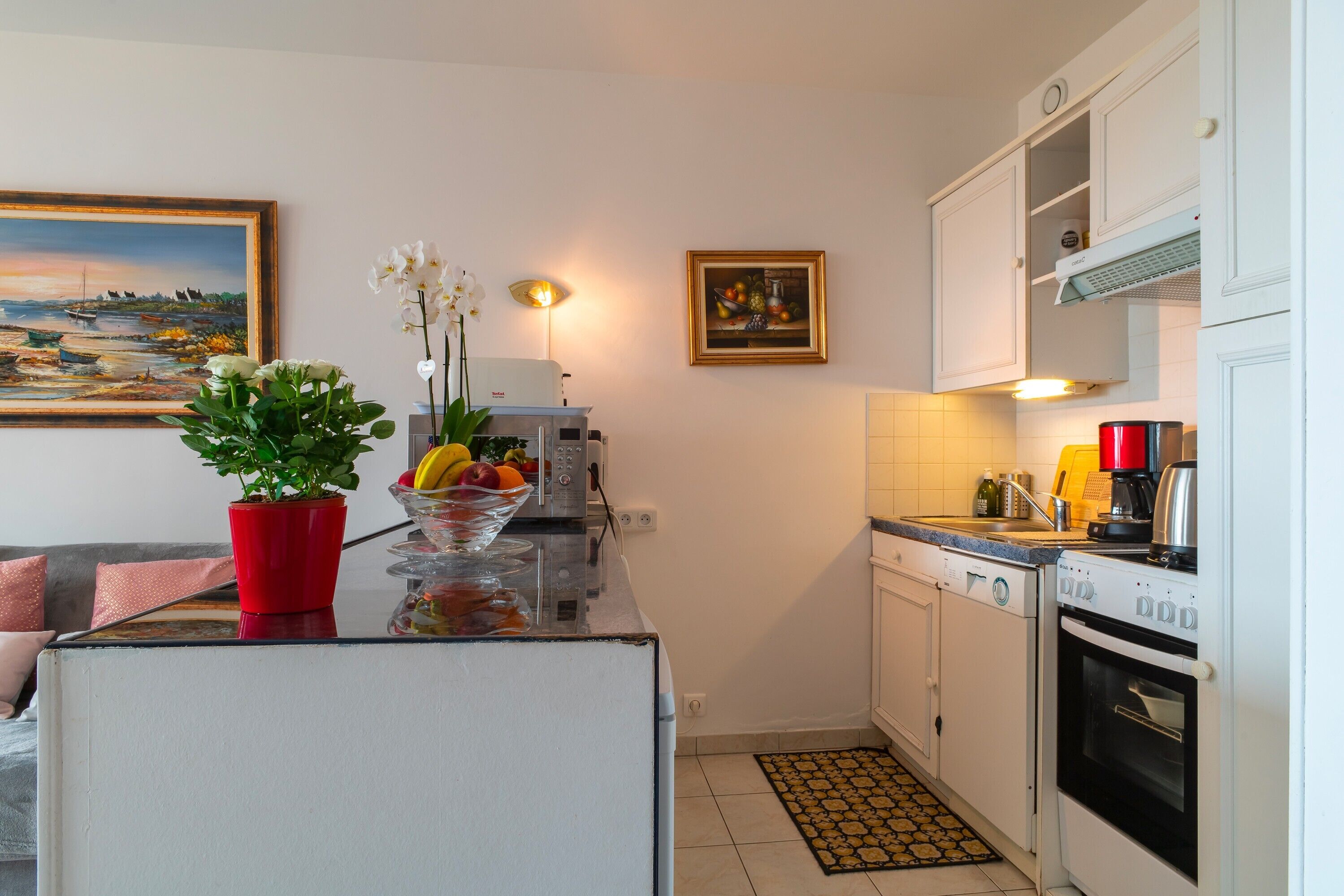 Une cuisine compacte avec des meubles blancs, un four encastré, une machine à café et une plante en pot posée sur le plan de travail.