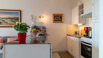 Une cuisine compacte avec des meubles blancs, un four encastré, une machine à café et une plante en pot posée sur le plan de travail.