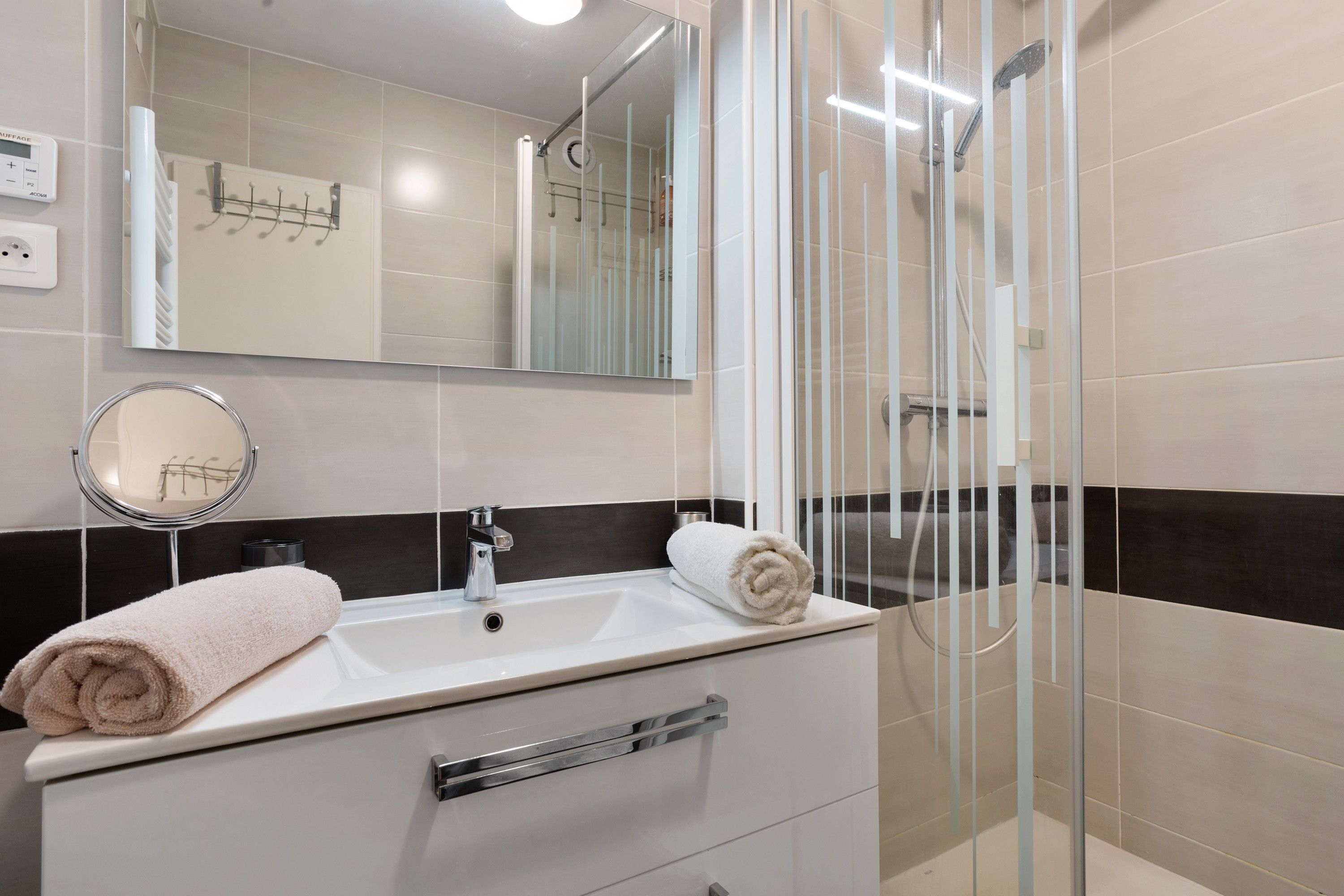 Ein modernes Badezimmer mit einem weißen Waschbecken, einem Spiegel, einem Handtuch und einer Duschkabine aus Glas.