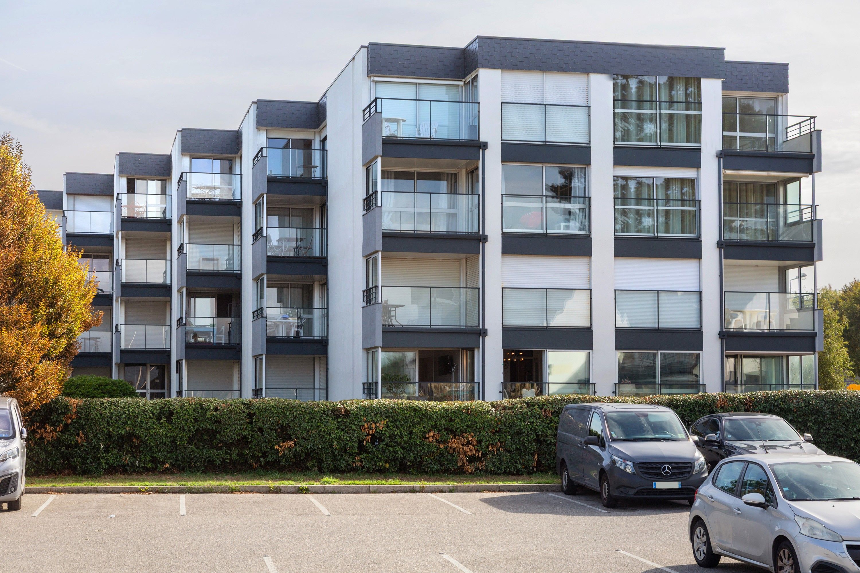 A multi-story residential building with balconies, a parking lot, and a hedge in front.