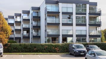 A multi-story residential building with balconies, a parking lot, and a hedge in front.