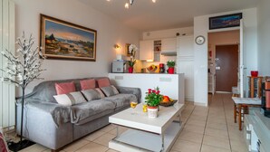 Una sala de estar con sofá, mesa de centro, un cuadro y un área de cocina con armarios y refrigerador.