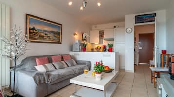 Un salon comprenant un canapé, une table basse, un tableau, et un coin cuisine avec des placards et un réfrigérateur.