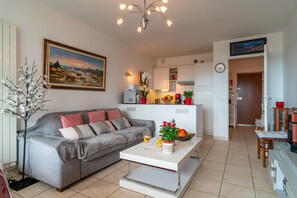 A living room with a sofa, a coffee table, a painting, and a kitchen area with cabinets and a refrigerator. - Apartment "Sea View" with Private Terrace, Balcony, and Wi-Fi (Bénodet)
