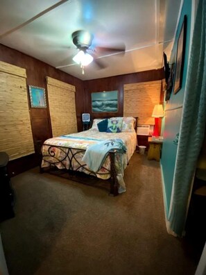 A bedroom with a bed, a ceiling fan, a nightstand with a lamp, and a wall-mounted TV. - Whimsical Mermaid Retreat with Private Deck in Bacliff, Texas (Bacliff)