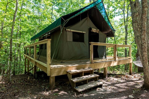 A wooden cabin with a green roof, a deck, and a window overlooking a forest.