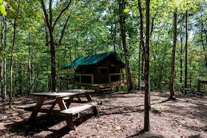Eine Holzhütte mit grüner Dachdeckung, umgeben von einem Wald. Im Vordergrund steht ein Picknicktisch.