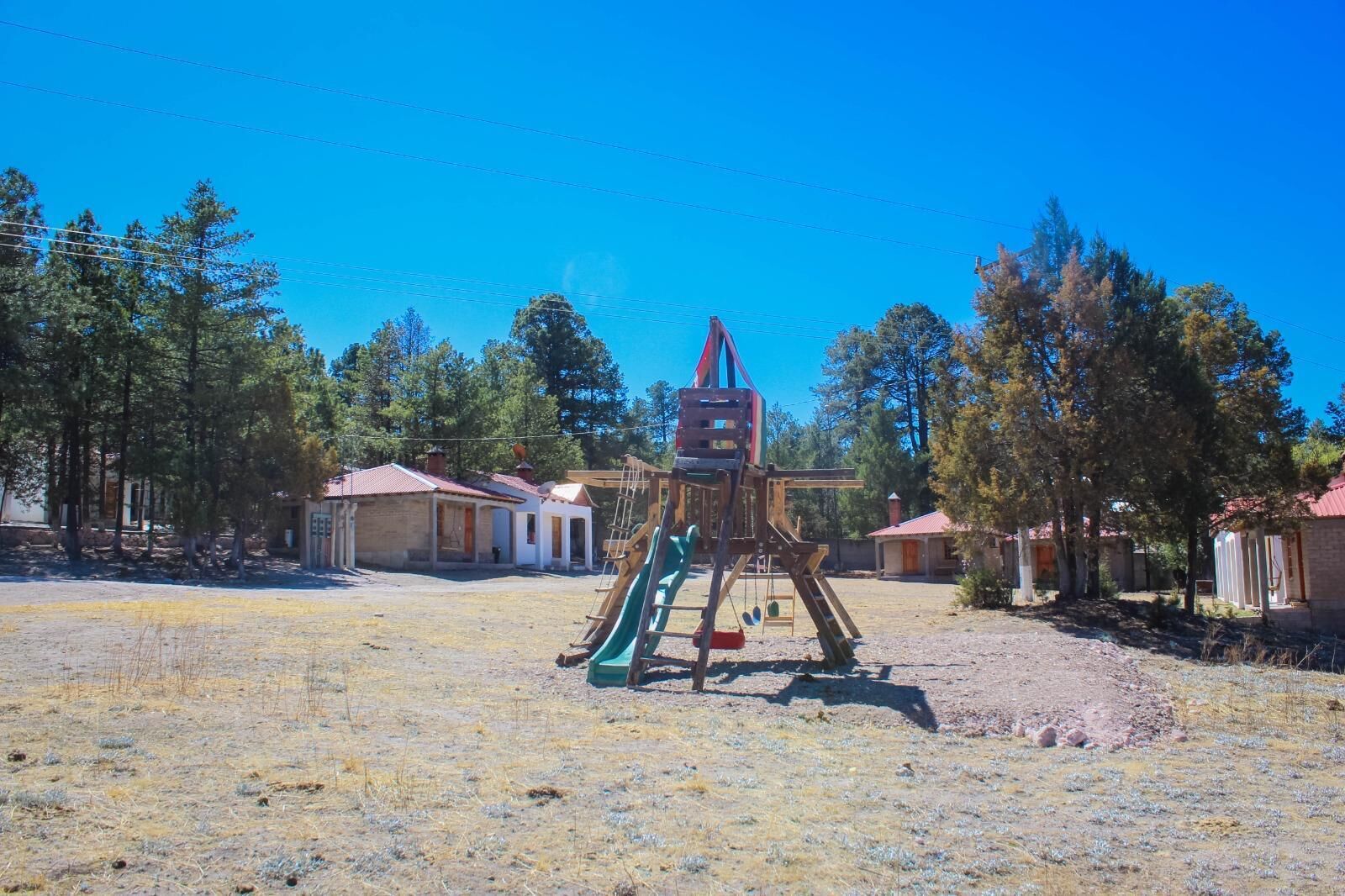A playground with a slide and climbing structure, surrounded by houses with red roofs, in a dry, grassy area with trees.