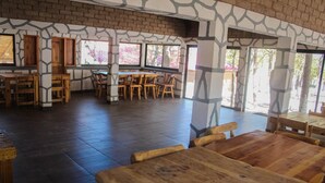 A spacious indoor area with wooden tables and chairs, large windows, and a brick wall with a unique pattern.