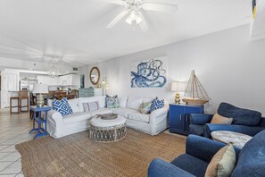 A living room with a white sofa, blue armchairs, a coffee table, and a dining area with a kitchen in the background. - 🌴Family Friendly Beach Condo | 5 Min Walk to Beach | Oceanwalk 4-404😍🐟🦈 (New Smyrna Beach)