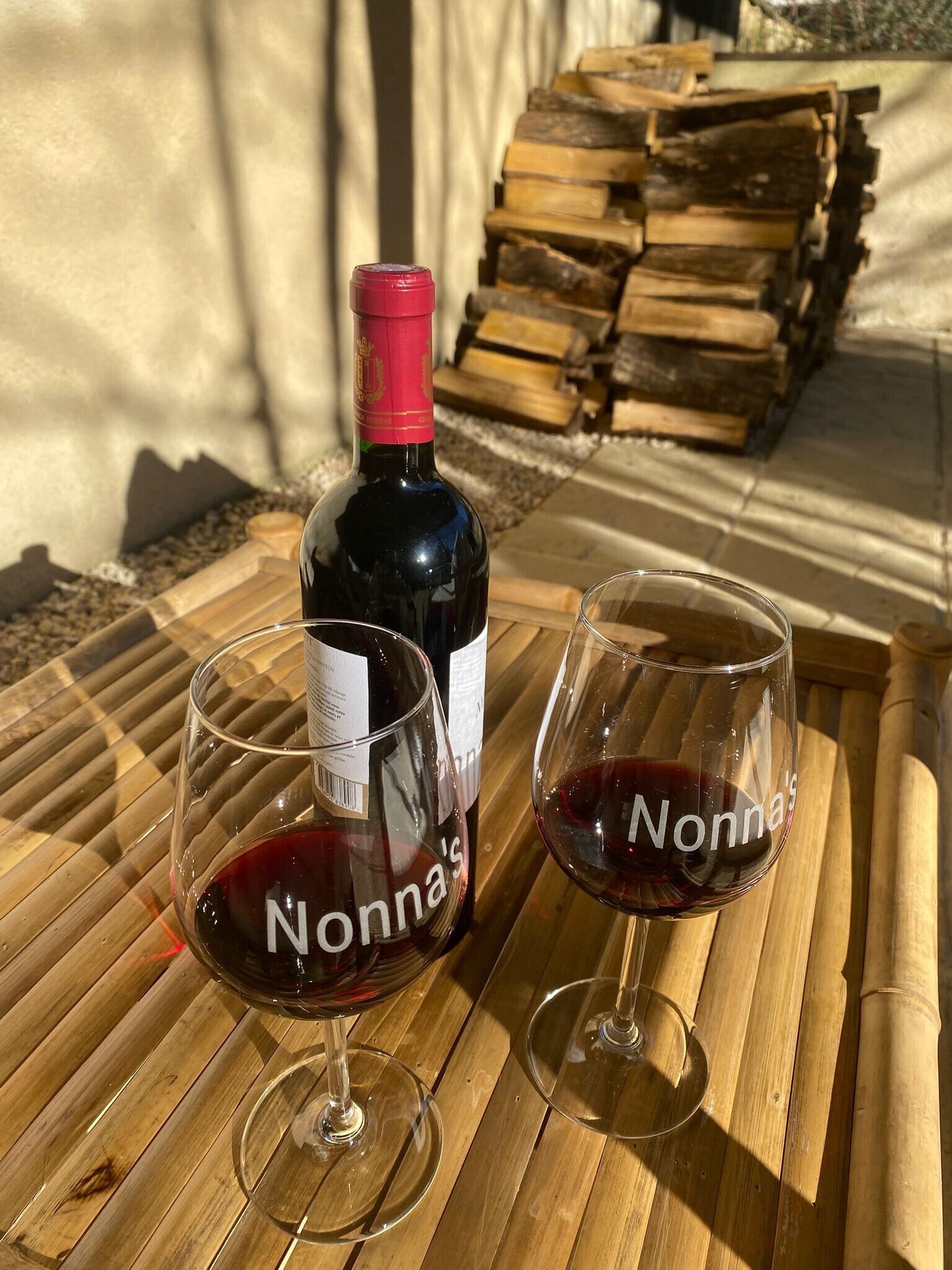 A bottle of wine and two glasses on a wooden surface with a stack of firewood in the background.