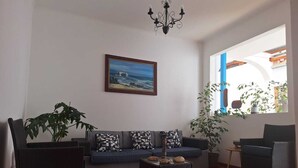 Una sala de estar con un sofá, dos sillones, una mesa de centro, una araña, un cuadro y una ventana con plantas.