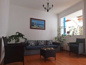 Ein Wohnzimmer mit einer Couch, zwei Sesseln, einem Couchtisch, einer Deckenleuchte, einem Gemälde und einem Fenster mit Pflanzen.