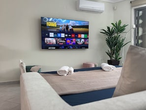 Ein Hotelzimmer mit großem, an der Wand montiertem Flachbildfernseher, der verschiedene Streaming-Dienste anzeigt, einem Bett mit ordentlich gefalteten Handtüchern, einer Topfpflanze und einer Klimaanlage.