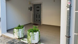 Entrada de un edificio con una puerta blanca, dos plantas en macetas y piso de baldosas.