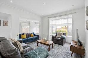 Ein geräumiges Wohnzimmer mit einem blauen Sofa, einem dunklen Sessel, einem Couchtisch und einem Fernseher auf einem Holzboden.