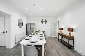Een eetkamer met een witte tafel gedekt voor zes personen, een grote koelkast, een klok en een console tafel met lampen.