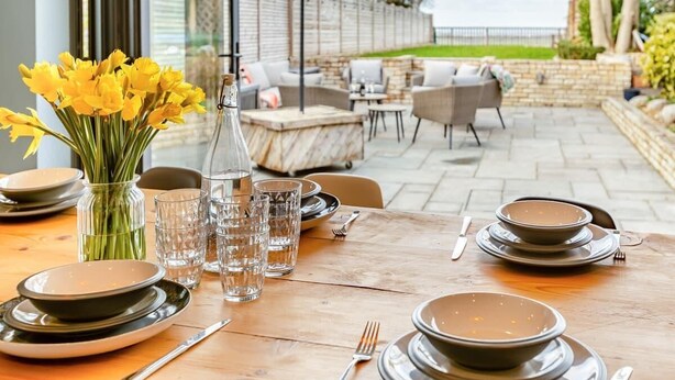Un espacio de comedor al aire libre con una mesa preparada para cuatro personas, un jarrón con flores amarillas y un muro de piedra al fondo.