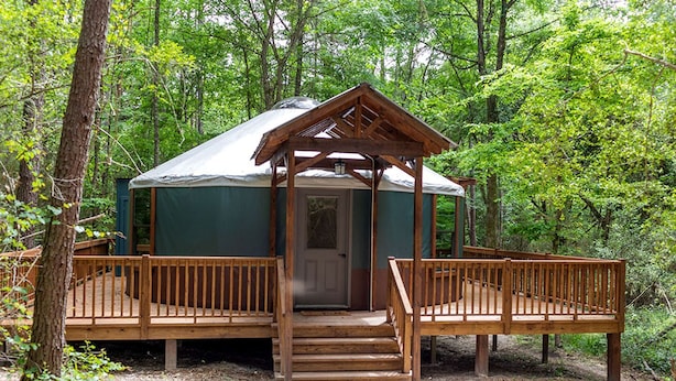 A wooden gazebo with a door, surrounded by a deck, set in a forested area. - Fully-Equipped Yurt Rental for Memorable Family Vacations in Livingston, Texas (Livingston)