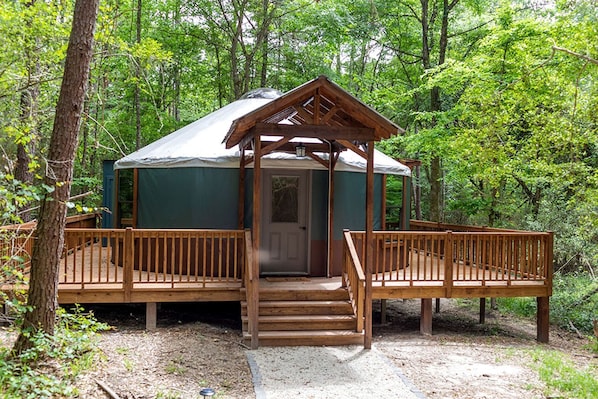 A wooden gazebo with a door, surrounded by a deck, set in a forested area.