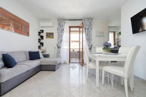 A modern living room with a sofa, a dining table with chairs, and a kitchen area visible through an open door.