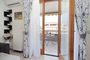 A room with a radiator, a potted plant, a small table with chairs, and a view of an outdoor balcony with a table and chairs.