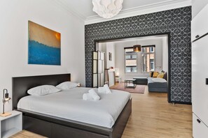 A modern bedroom with a large bed, a painting, and a view into the living area.