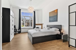 Ein modernes Schlafzimmer mit einem großen Bett, einem Nachttisch mit Lampe, Blick auf die Stadt durch ein Fenster und einem Kleiderschrank.