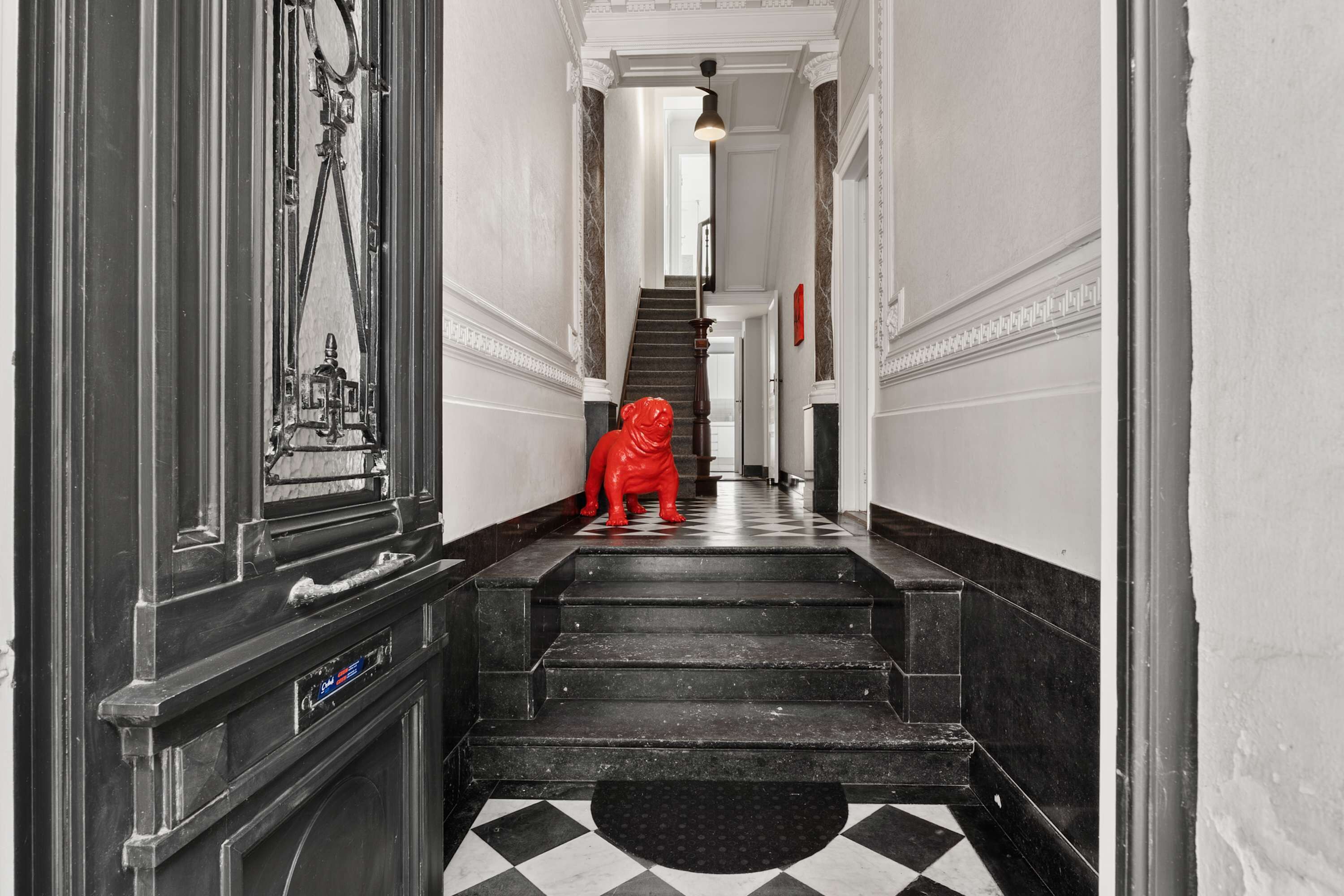 Un couloir doté d’un sol à carreaux noirs et blancs, d’une statue rouge et d’un escalier menant à une porte ornée d’une fenêtre circulaire.