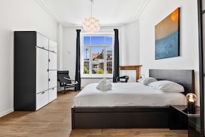 Une chambre d’hôtel moderne équipée d’un lit, d’une chaise, d’un bureau, d’une armoire et d’une fenêtre donnant sur des immeubles.